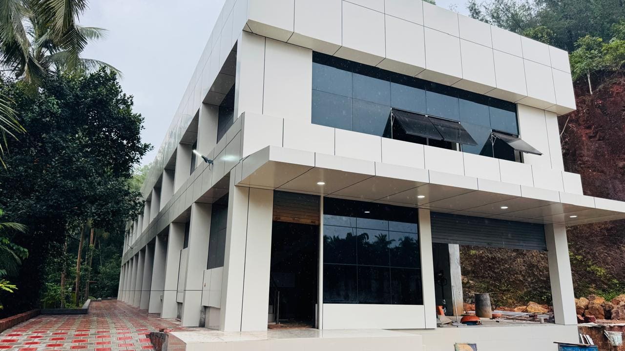 The two-storey white composite-clad facility building at Kudukoli, against a laterite cutting and palms.